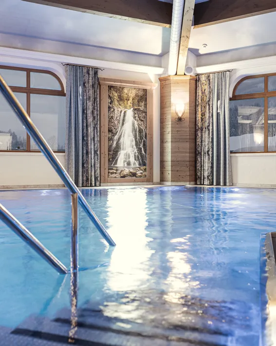 Hotelhallenbad mit warmer Beleuchtung, Wasserfallbild und Bergblick durch große Fenster